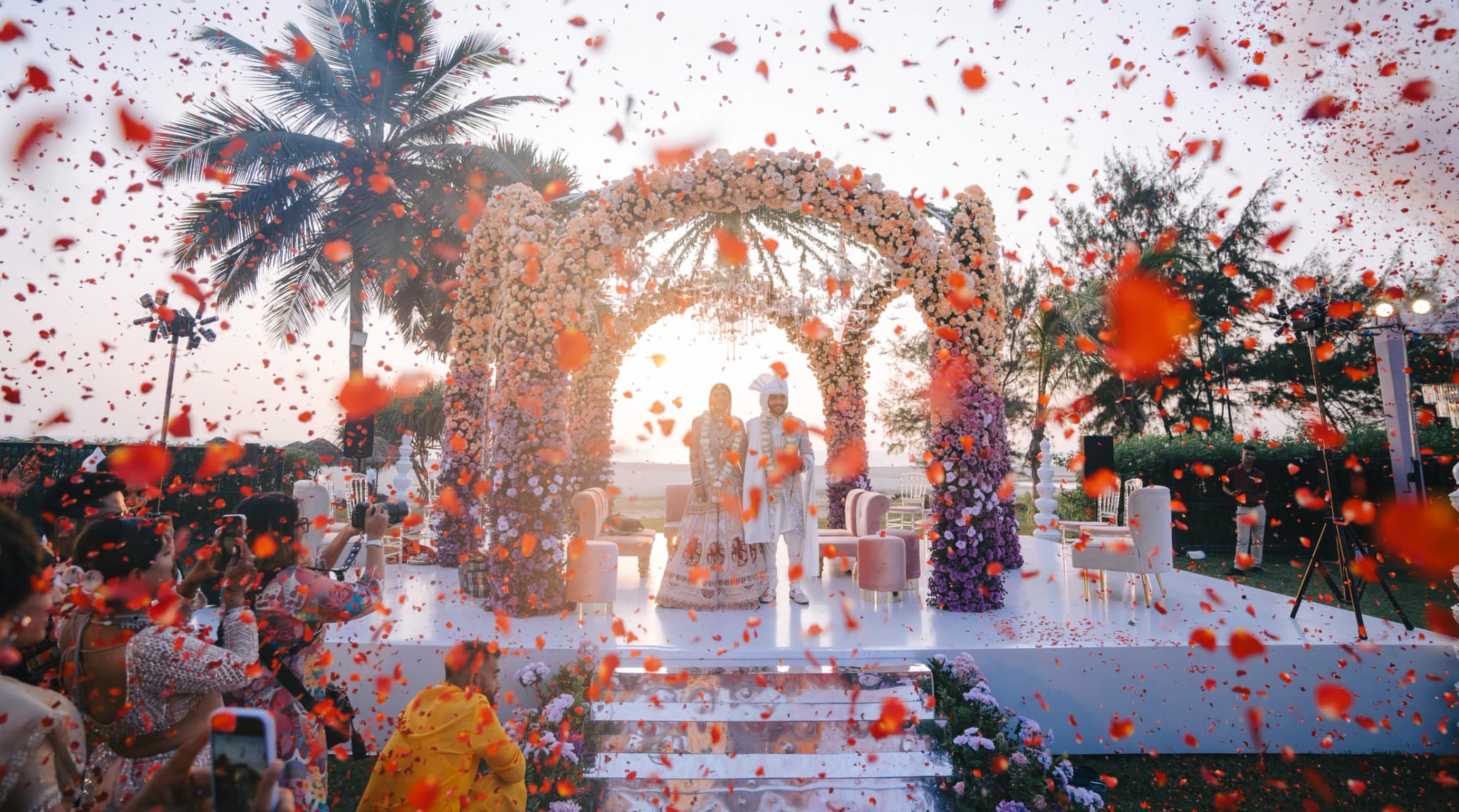 Wedding couple beneath a floral ceremony arch surrounded by falling red petals