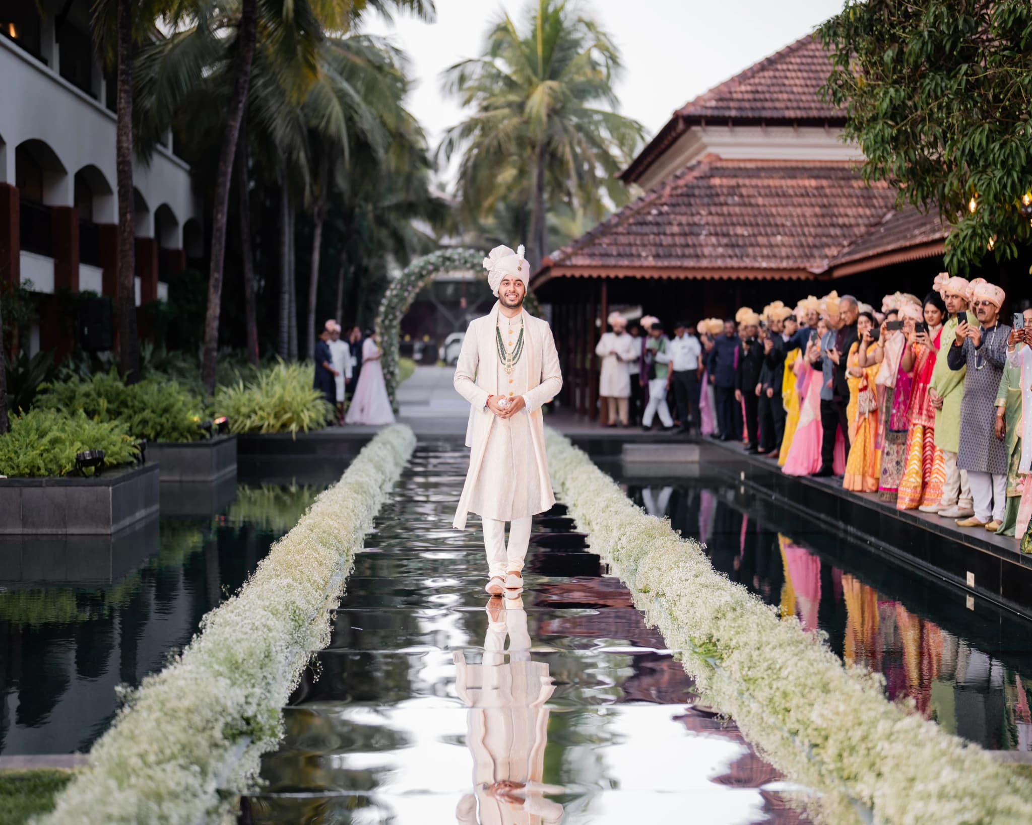 Groom walking along a mirrored wedding aisle lined with white flowers and guests