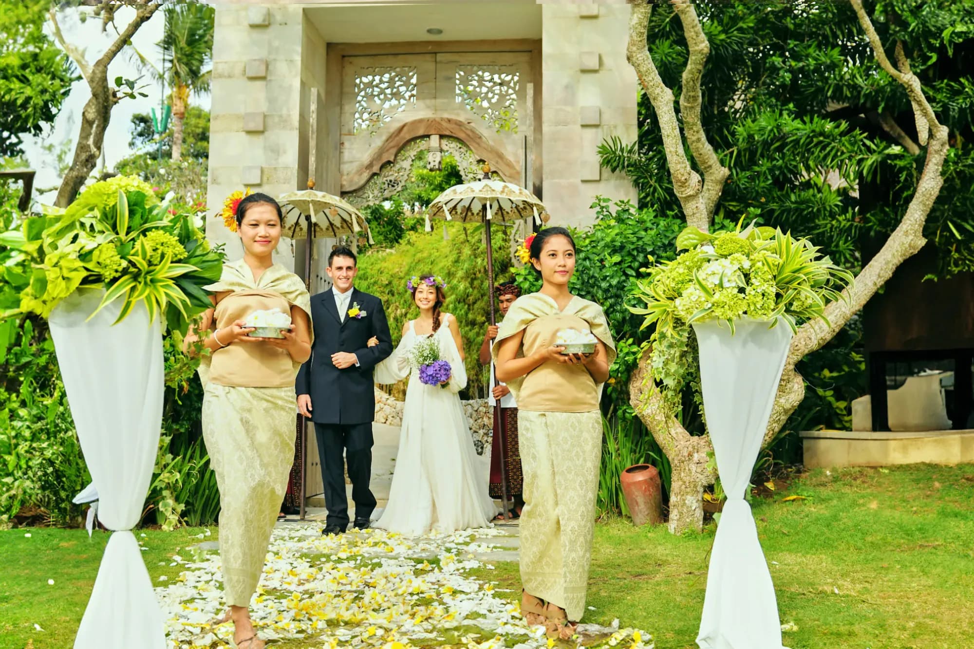 Balinese candi bentar split gate with attendants holding flower trays and frangipani petals at AYANA Estate Bali