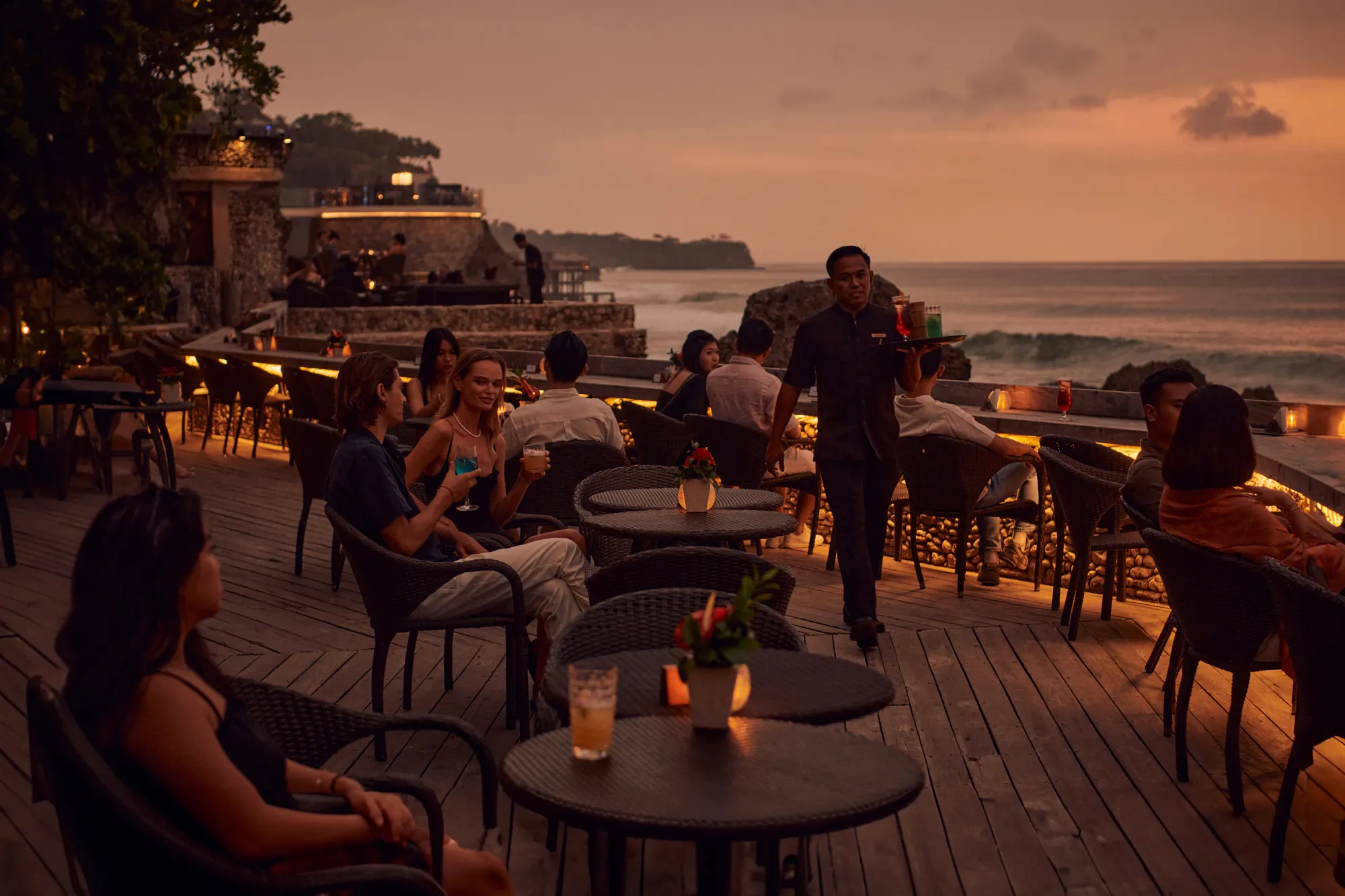 Guests at tables on the Rock Bar deck over the ocean at sunset at AYANA Estate Bali, Jimbaran