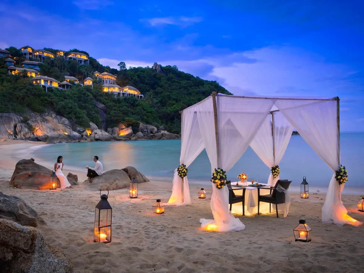 Draped wedding canopy with lanterns on the private beach at twilight below the hillside villas at Banyan Tree Samui
