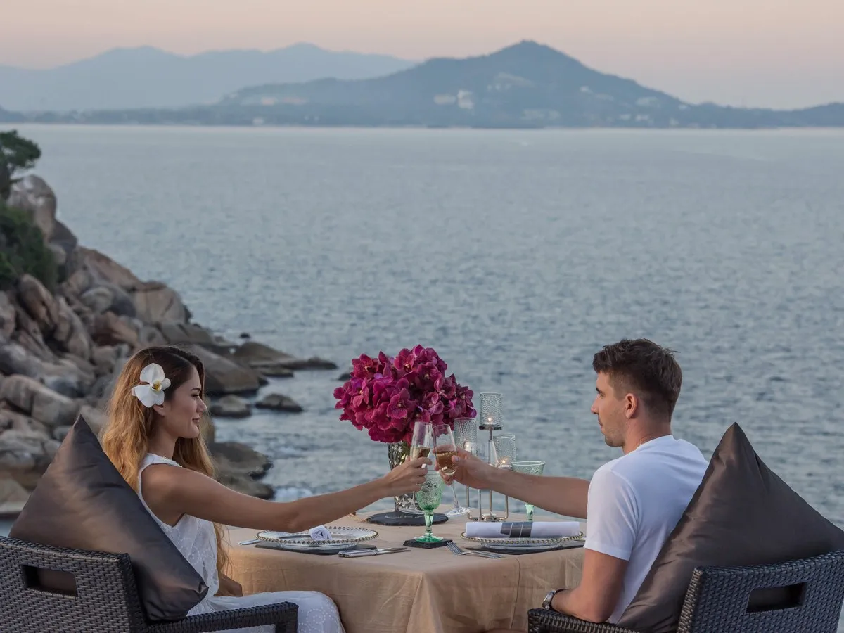 Couple toasting at a candle-lit clifftop table overlooking the Gulf of Thailand at sunset, Banyan Tree Samui