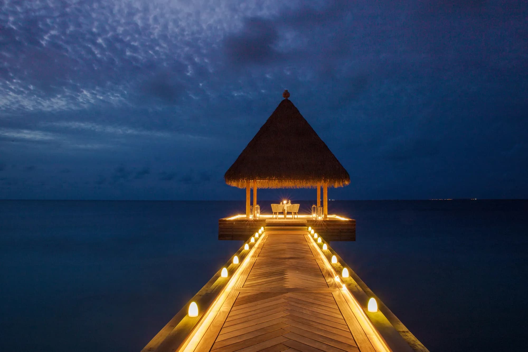 Over-water pavilion lit with candles along the jetty at blue hour at Waldorf Astoria Maldives Ithaafushi