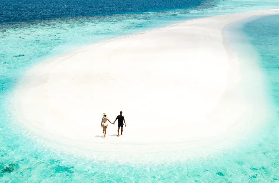 A couple walking hand in hand on a white sandbank surrounded by turquoise water at Waldorf Astoria Maldives Ithaafushi