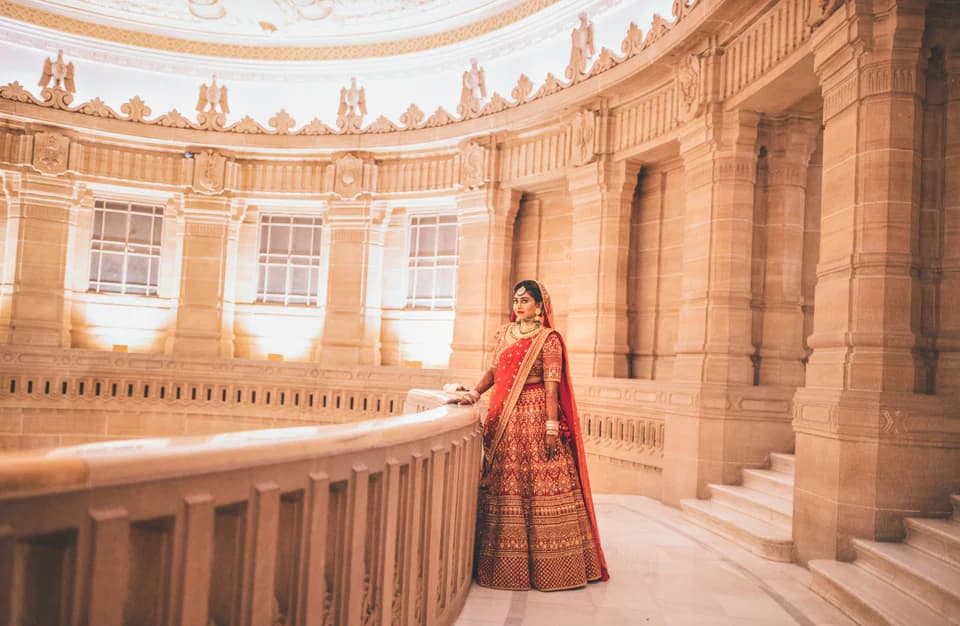 Bride in a red lehenga standing along a grand sandstone palace colonnade