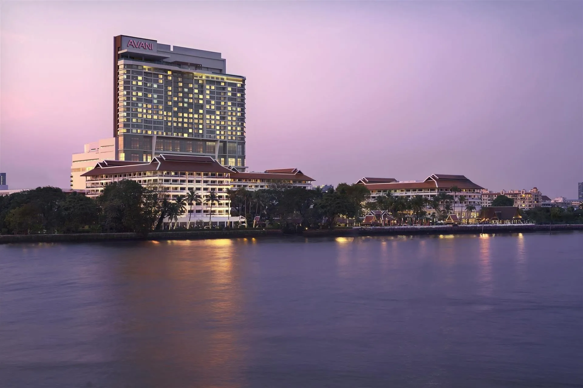 AVANI+ Riverside Bangkok high-rise hotel tower glowing above the Chao Phraya River at dusk, Bangkok