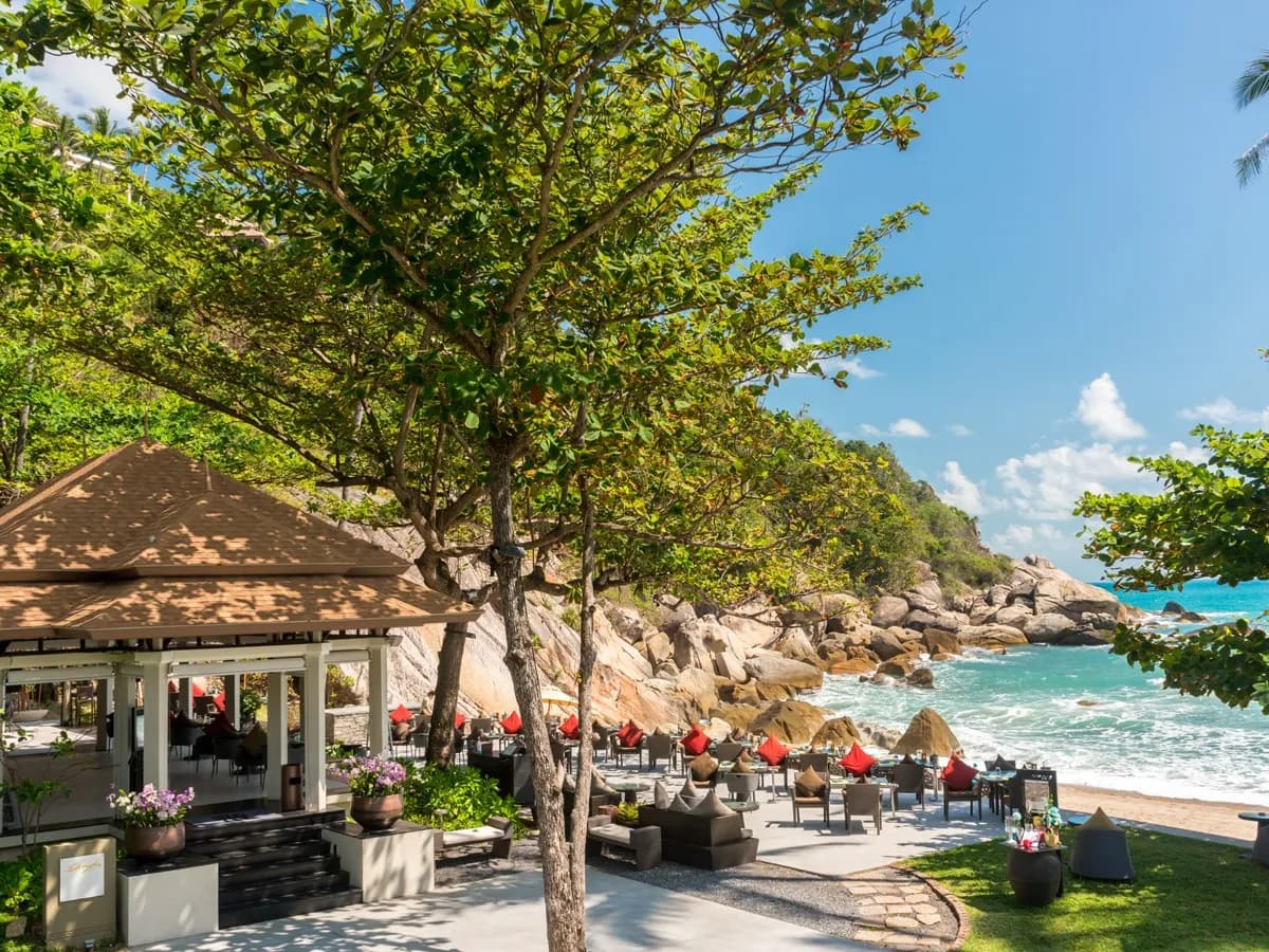 Beachfront pavilion lounge framed by trees beside the rocky private cove at Banyan Tree Samui, Koh Samui