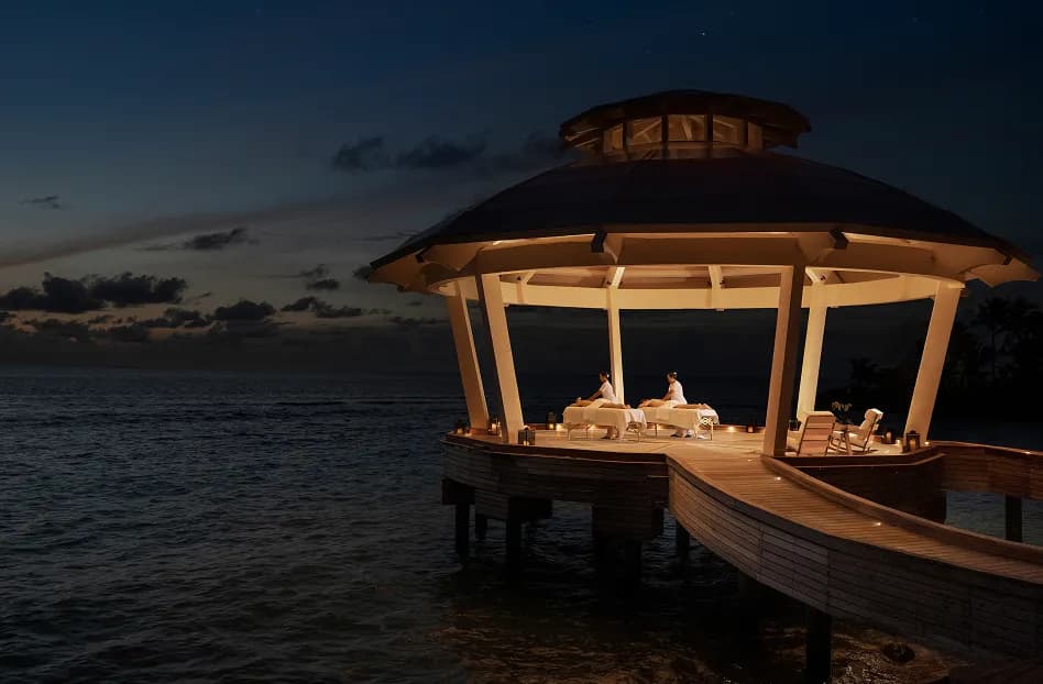 Thatched over-water pavilion lit with lanterns along the jetty against the Indian Ocean at dusk, Waldorf Astoria Maldives Ithaafushi